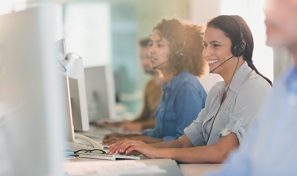Call center workers wearing headsets are sitting along a desk, providing customer support over the phone.