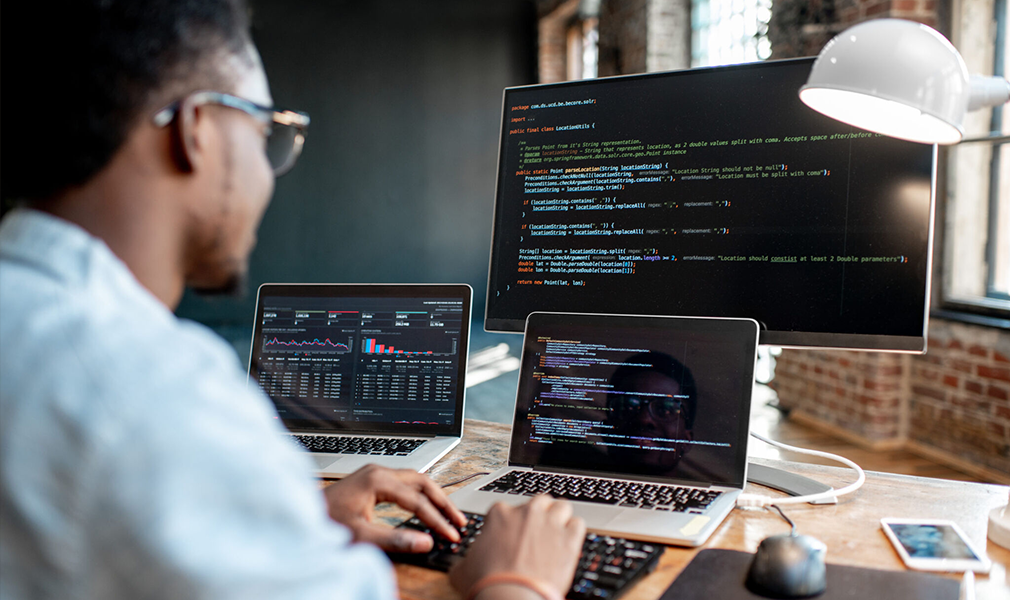 A software developer sits at a desk and looks at 3 screens with code on them.