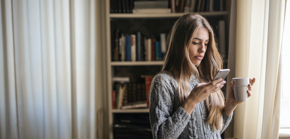 A person is holding a cup of coffee in one hand, and looking at their smartphone in the other hand.