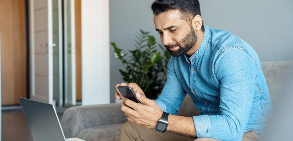 A person is sitting on the couch in front of their computer, holding and looking at a smartphone.