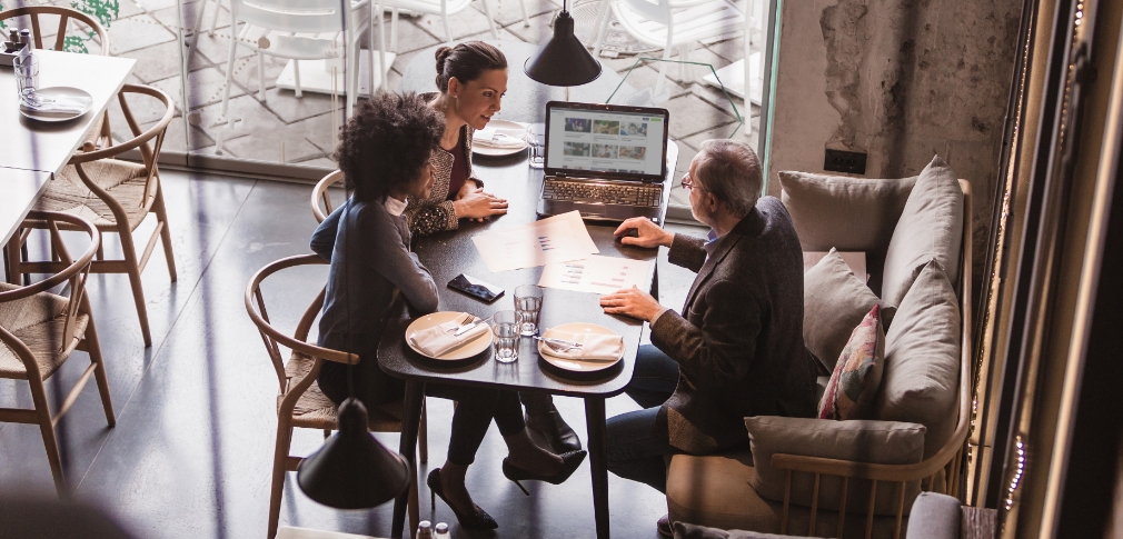 Three people sitting around a table at a cafe, and looking at an open laptop on the table.