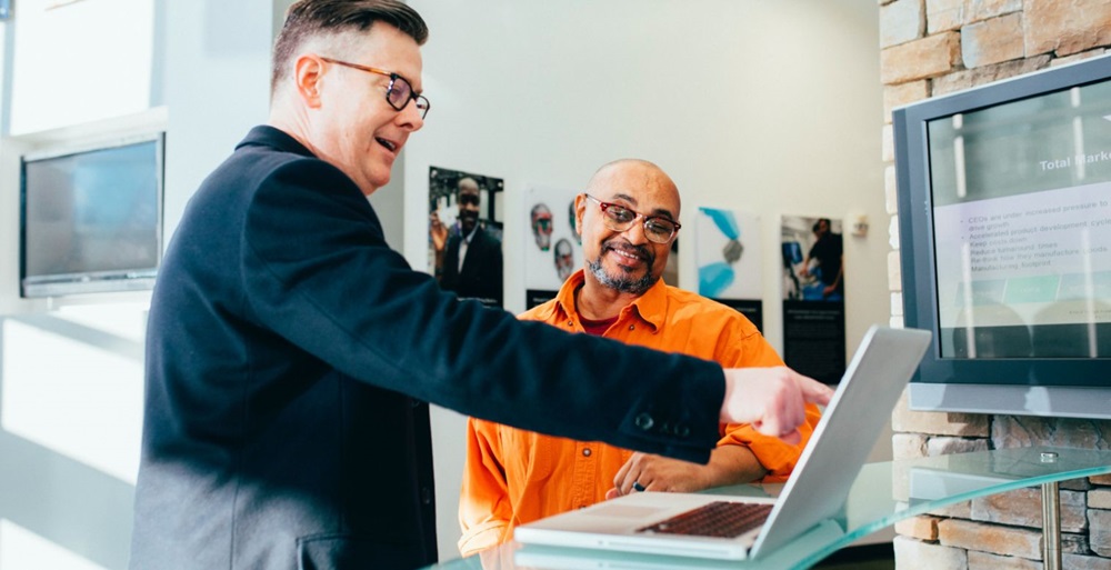 Two people standing around a laptop and pointing at it.