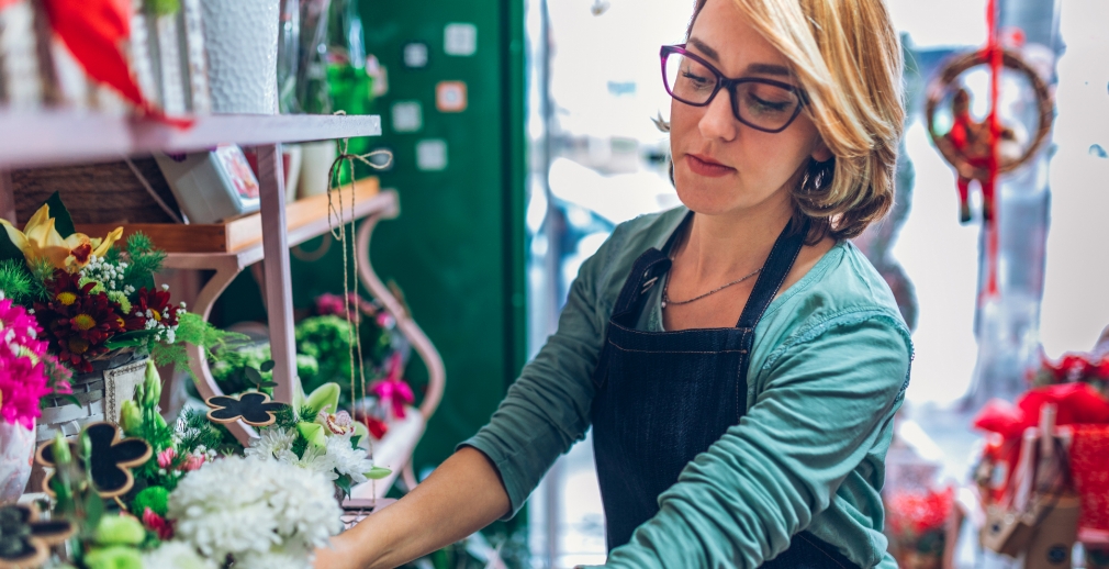 Woman with blond hair wearing glasses, a green shirt and black apron merchandising in a plant store, with holiday decorations in the background