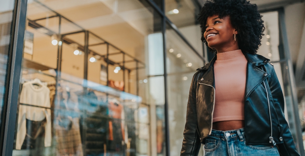Woman wearing a pink shirt and black leather jacket smiling while window shopping outside a clothing boutique