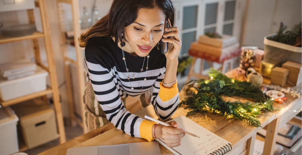 Woman talking on phone and taking handwritten notes in a retail store, surrounded by unpacked holiday decor