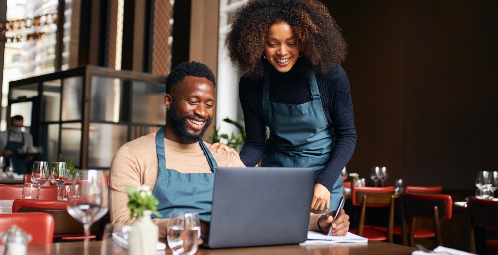One male and one female restaurant employee wearing teal aprons looking at a laptop and laughing