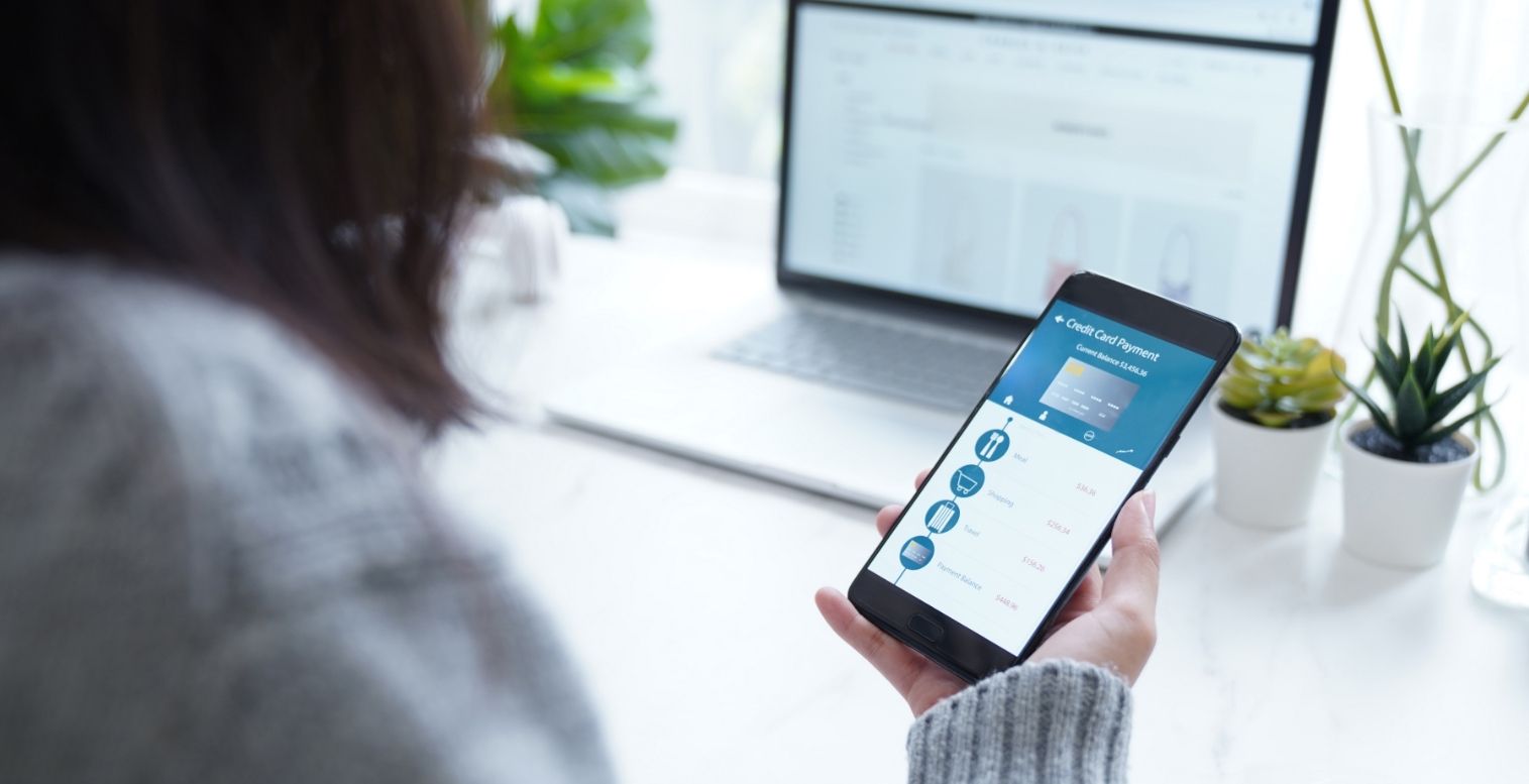 Woman sitting at desk looks down at smartphone to view her credit card balance and banking information