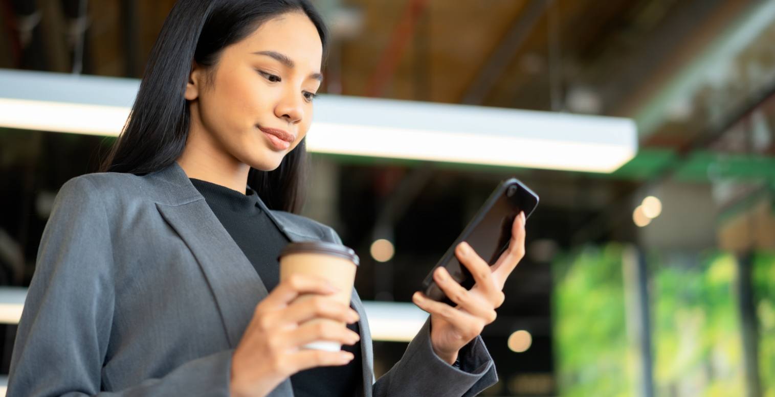 Woman in gray blazer holds a coffee cup and unlocks her smartphone with biometric facial recognition technology