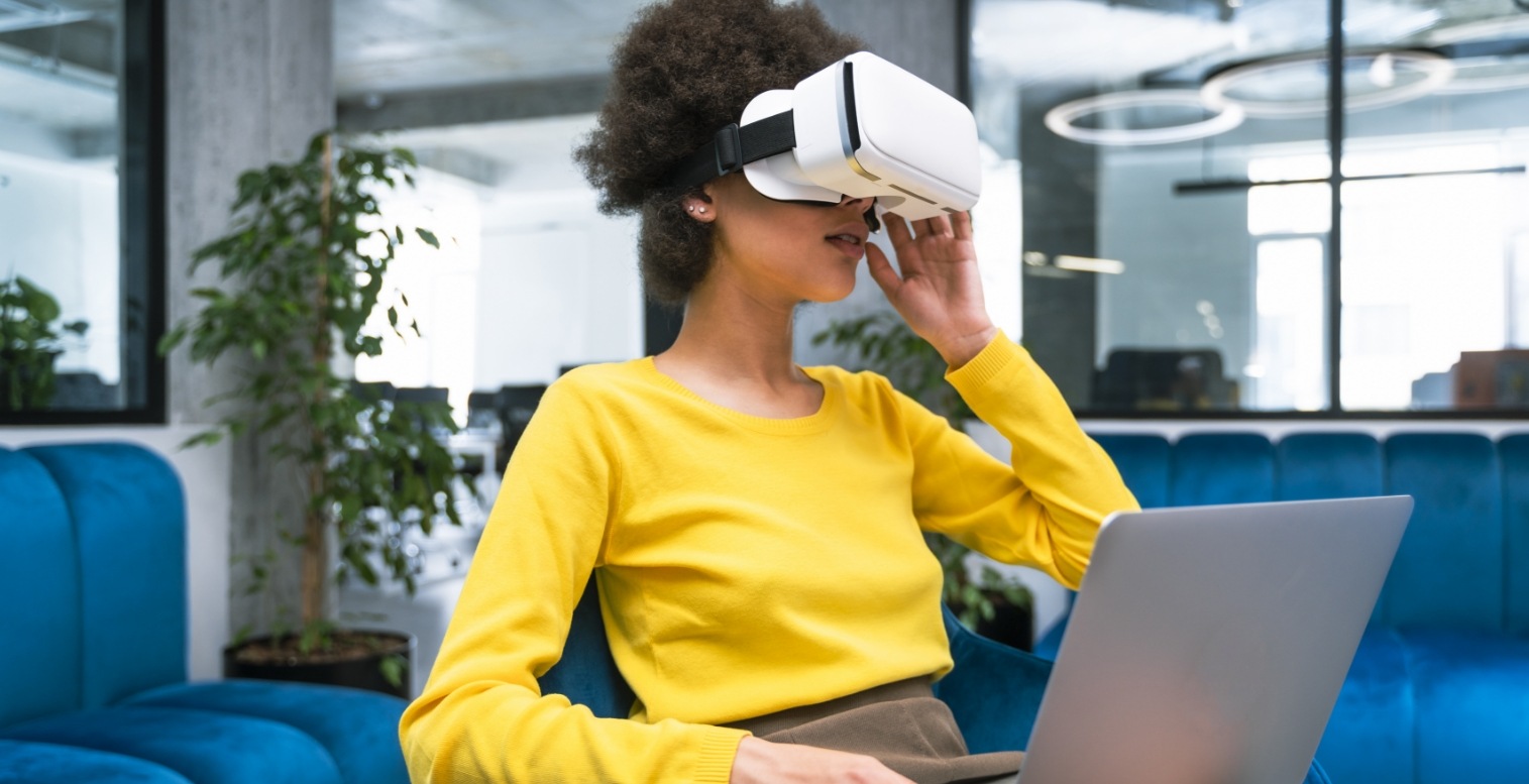 Woman in yellow shirt wears a virtual reality headset while holding her laptop on her lap