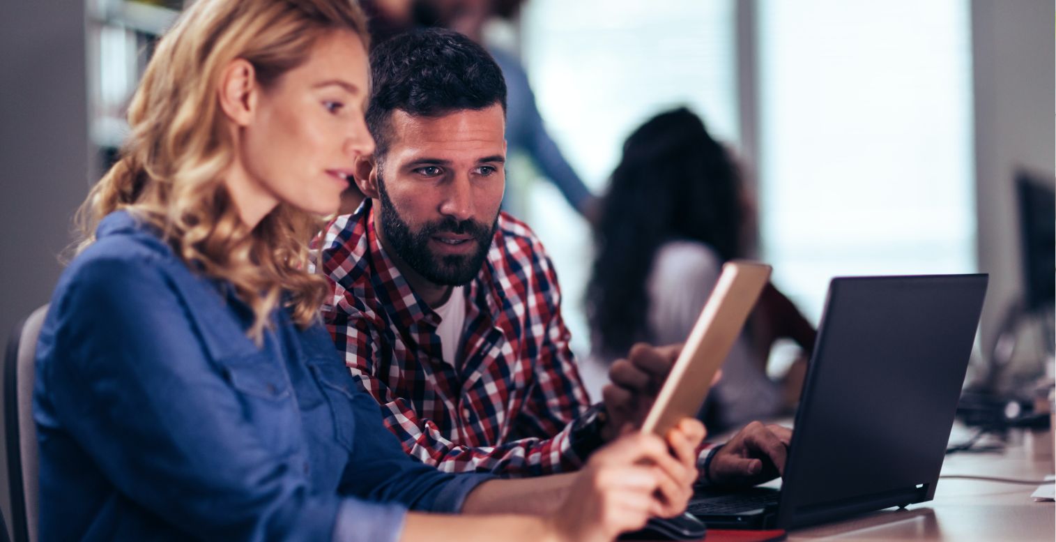 Woman in blue shirt shows man in plaid shirt information on her tablet about payroll and HR software.