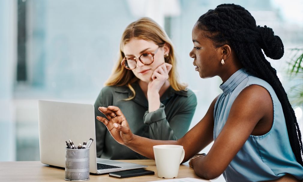 Two women in an office setting look at a laptop together.