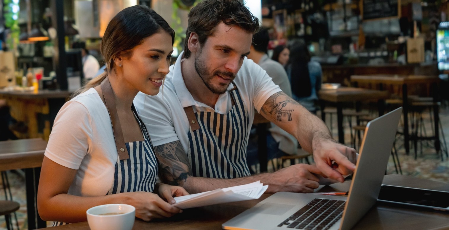 Two restaurant employees sit at a table and look at a laptop together.