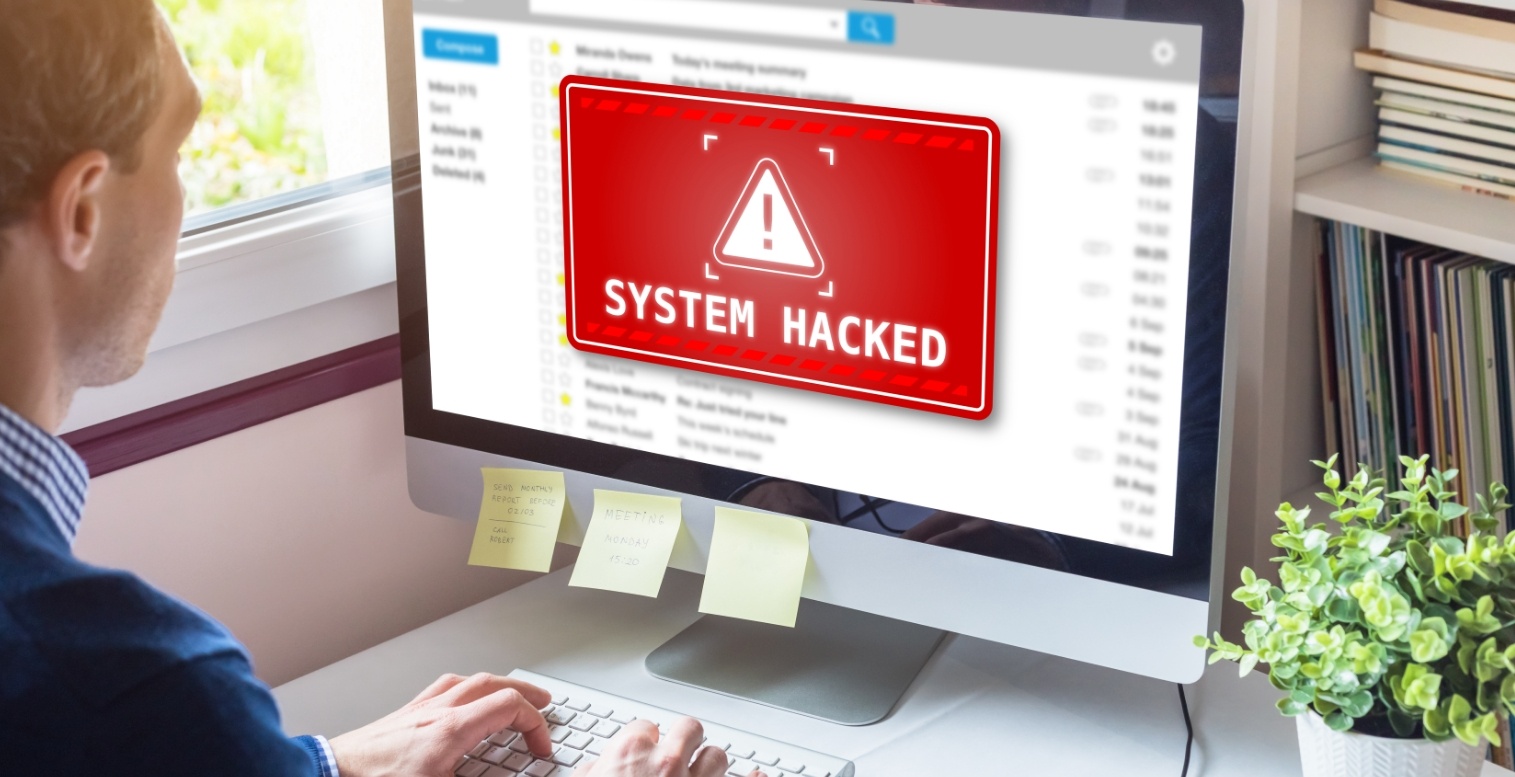 A man sits at a computer. The screen displays a large warning that reads “System Hacked.”