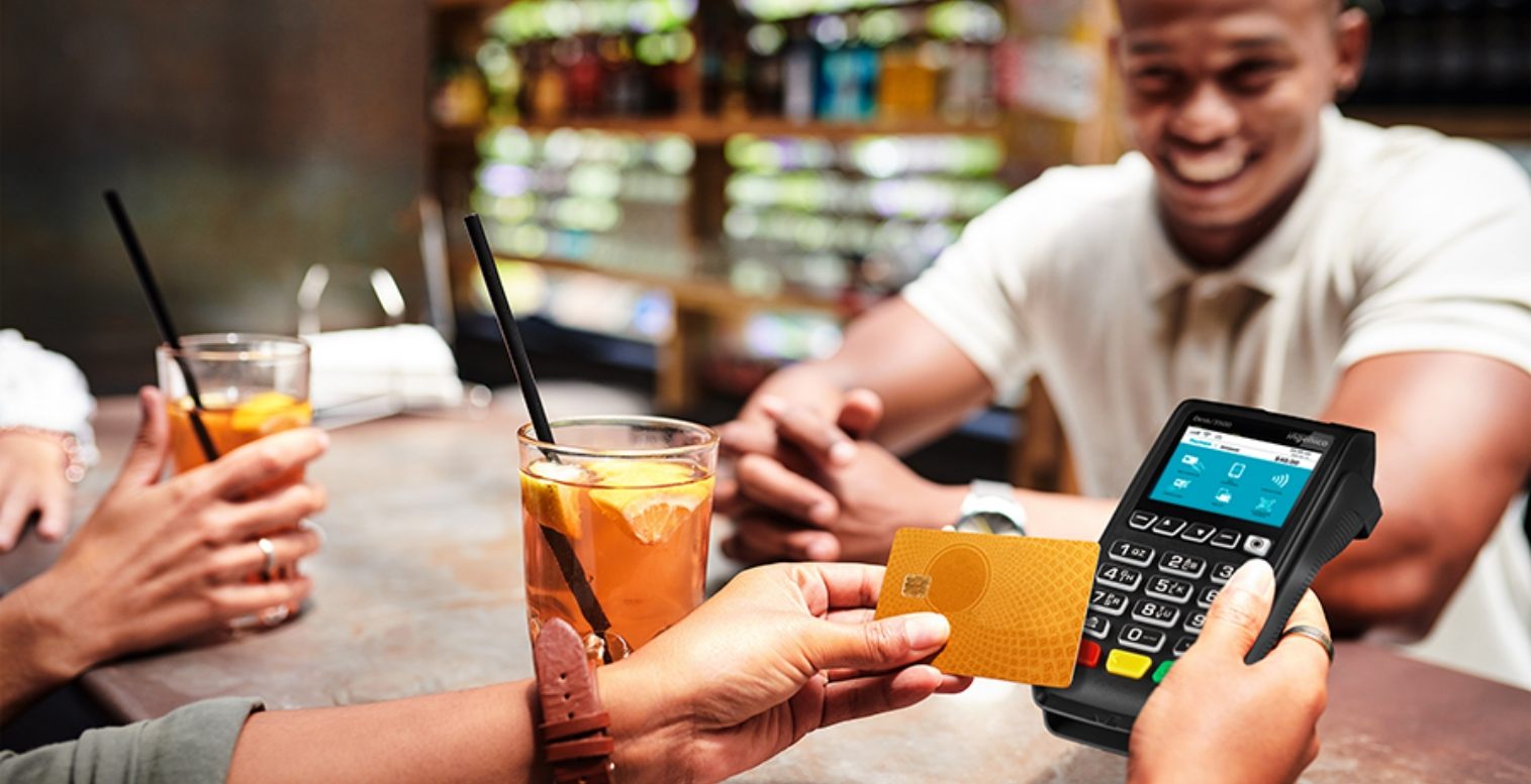 A group sits at a restaurant table, and one patron uses their credit card to pay on a mobile point of sale device.