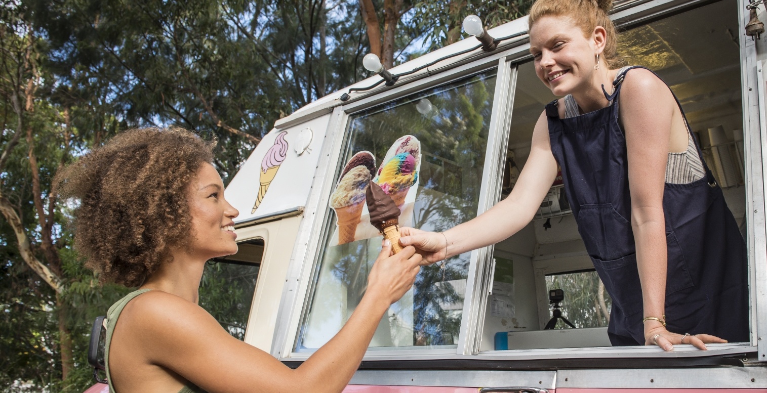 A happy woman wearing an apron hands another smiling woman an icecream cone from the window of a food truck.