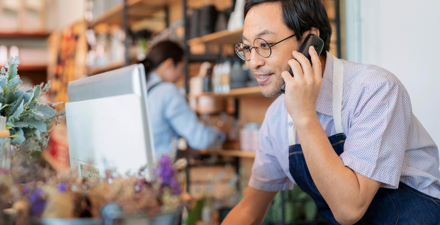A male wearing an apron and glasses leans close to a desktop computer screen and speaks on a handheld phone in a retail shop while a female employee moves around items on a shelf in the blurry background.