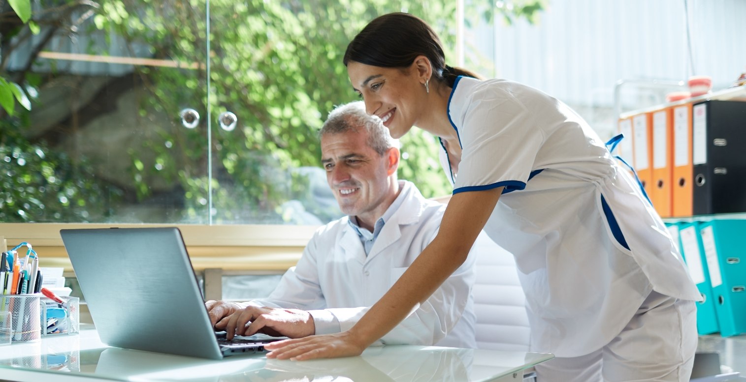 A male dental office employee sits at his desk while a standing female employee leans over to help him with a task on his open laptop.