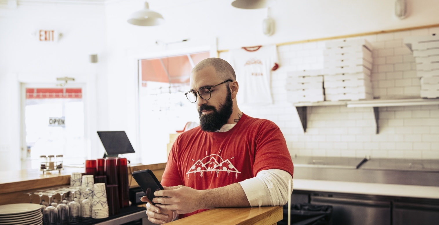 A male worker wearing glasses at a pizza restaurant looks down at a tablet he’s holding while standing behind the food preparation counter.