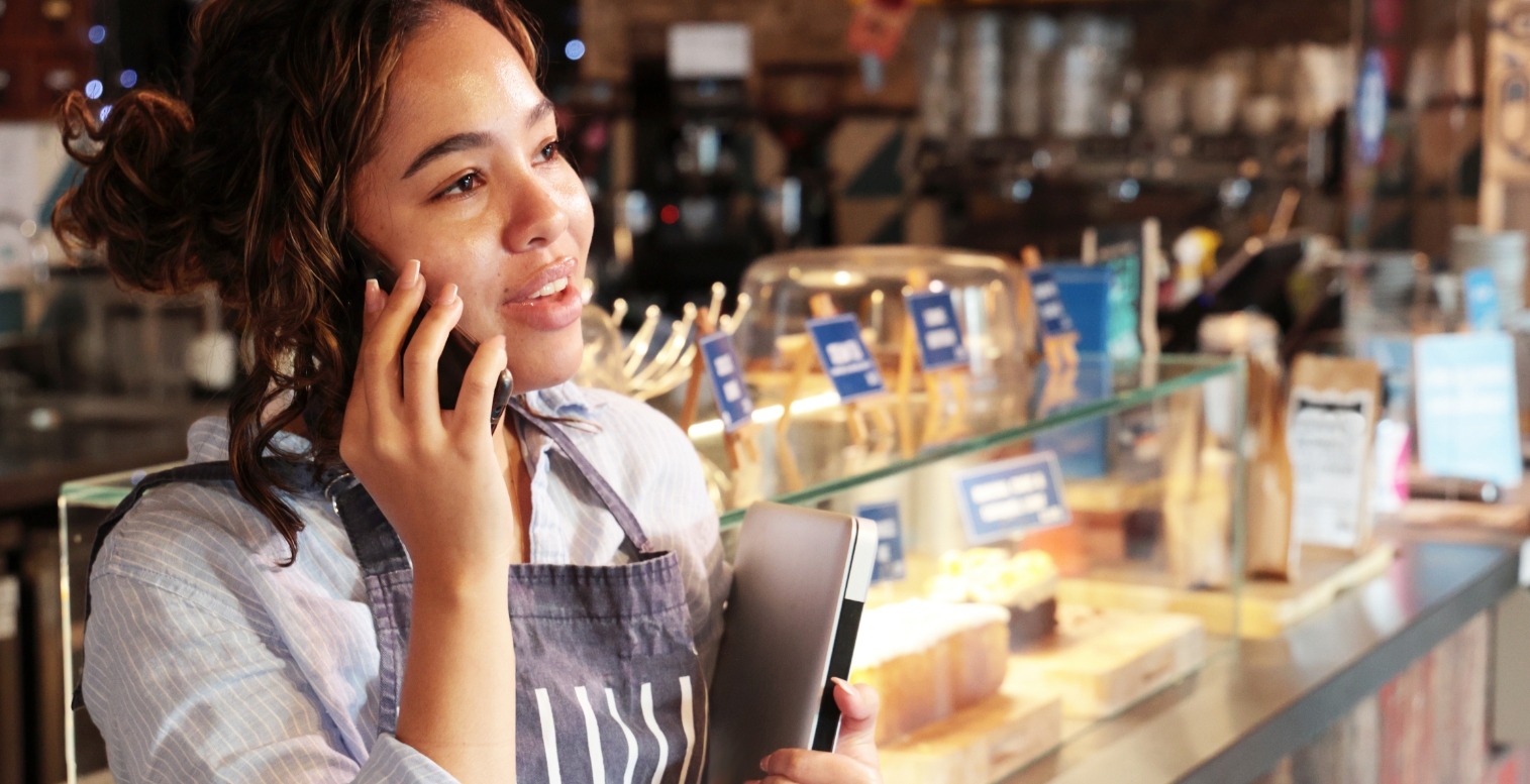 A woman wearing an apron and holding a closed laptop stands in front of a cafe counter while speaking on a handheld phone.