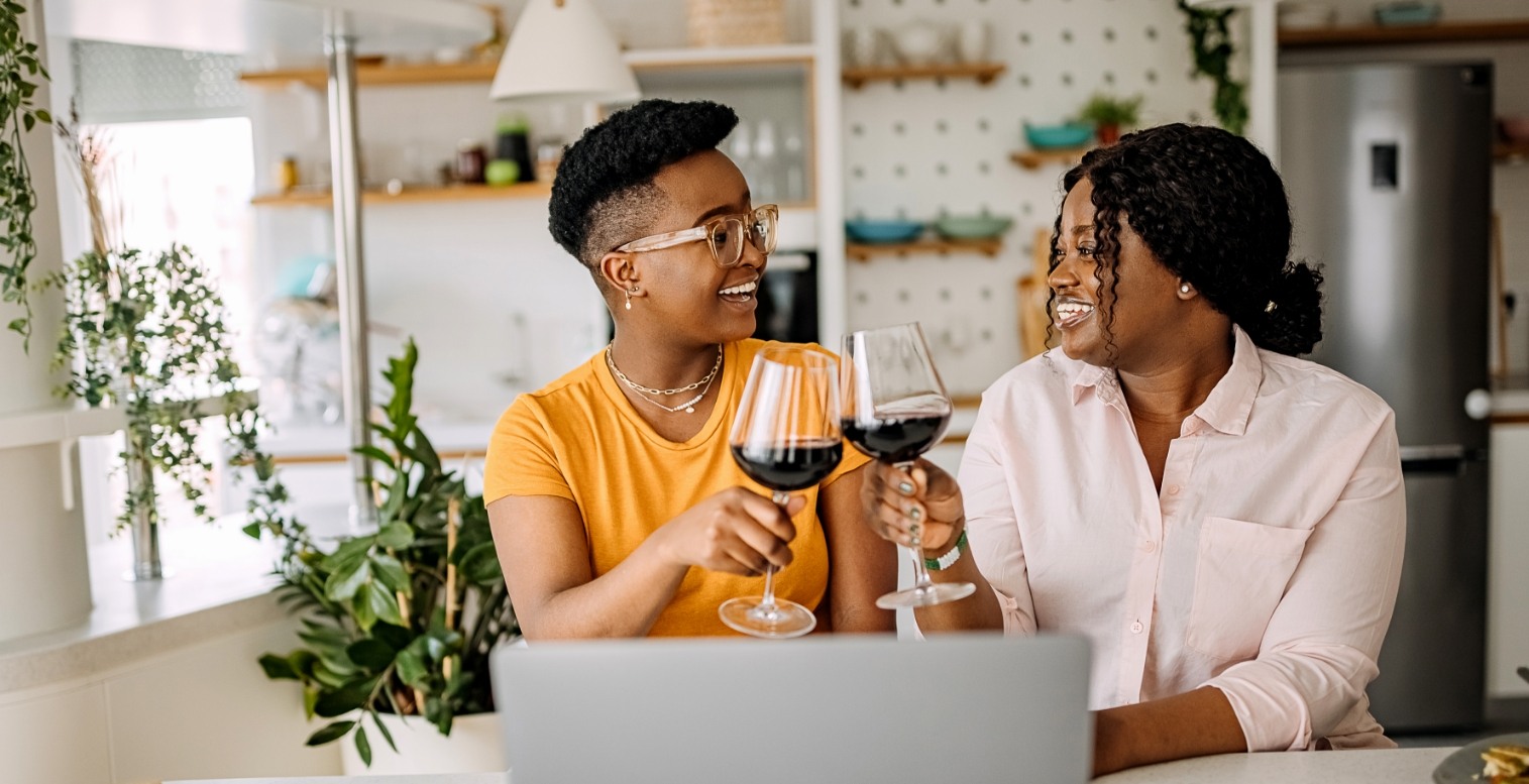 Two female employees in casual clothing, one wearing glasses, cheers their glasses of red wine while sitting in front of an open laptop at a counter.