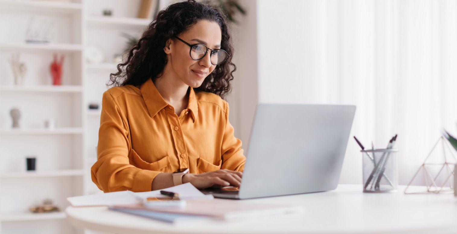 A woman wearing glasses sits at a desk and looks at her open laptop.
