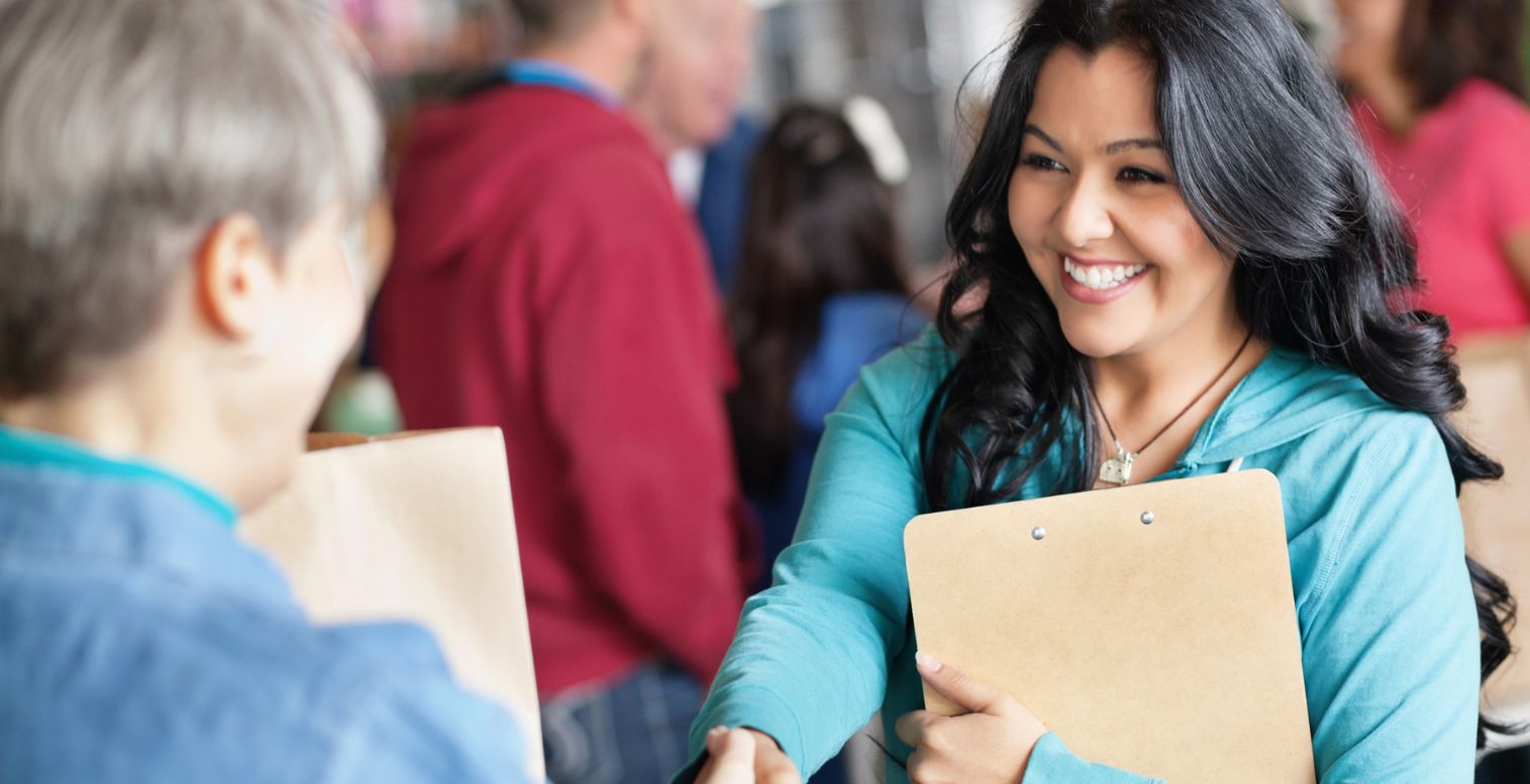 A smiling woman with a clipboard shakes hands with another person with a clipboard whose back is turned.
